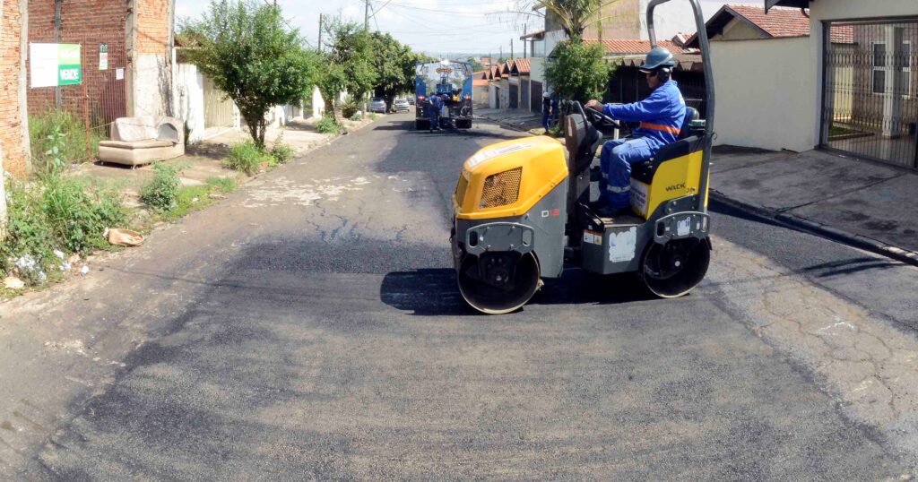 Um rolo compactador passa sobre o novo asfalto para nivelar e compactar a via, enquanto a equipe de manutenção atua ao fundo da rua.