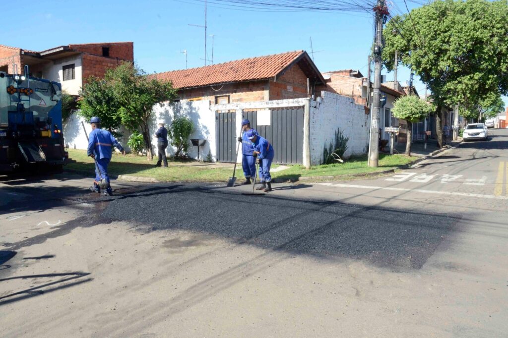 Na imagem, vemos uma cena de manutenção urbana em um bairro residencial: vários trabalhadores com uniformes azuis e capacetes de segurança estão espalhando e nivelando uma nova camada de asfalto com pás e ancinhos, enquanto um grande veículo de manutenção viária auxilia no processo de compactação; ao fundo, aparecem casas de telhados vermelhos e paredes brancas cercadas por árvores e arbustos, e no chão destaca-se a palavra “PARE” pintada em branco, compondo um retrato do cotidiano de reparos que garantem melhores condições de tráfego e segurança.