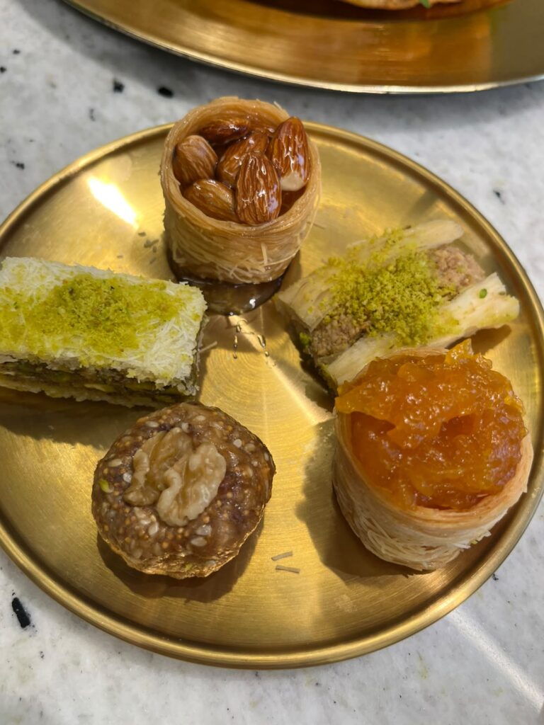O prato mostra uma seleção refinada de doces árabes servidos em um prato dourado, criando um contraste elegante com a mesa de mármore clara.