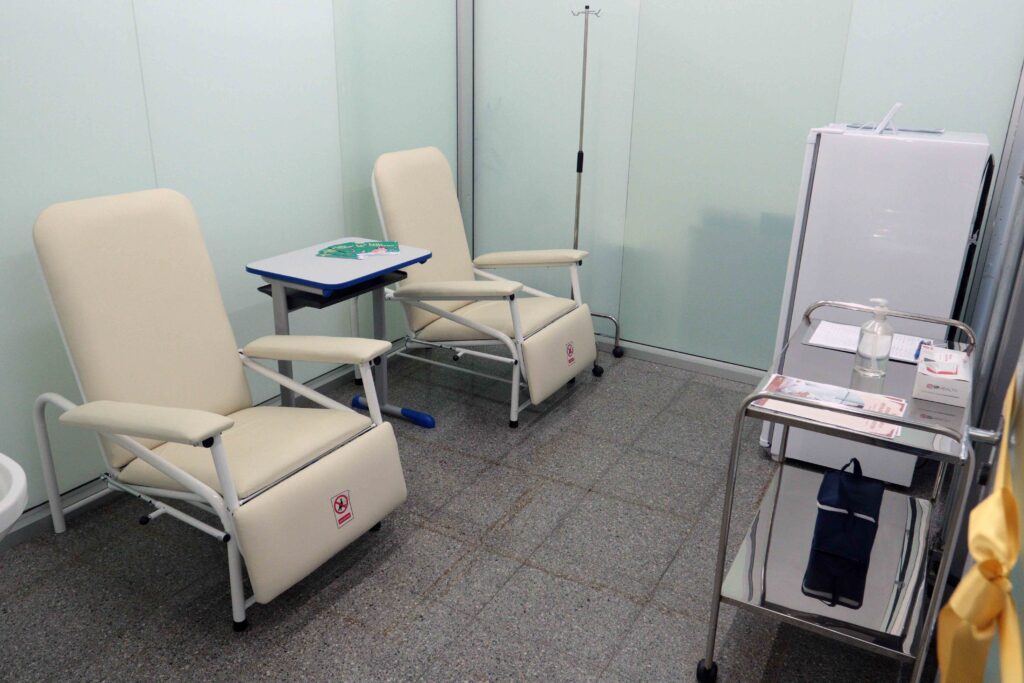 A imagem está girada e mostra uma sala de aula ou treinamento, com várias carteiras escolares brancas e azuis organizadas em fileiras. Sobre as mesas há apostilas, folhas impressas, canetas e outros materiais. O ambiente tem paredes claras, piso cinza, cadeiras com pranchetas acopladas e algumas placas de segurança nas laterais. Não há pessoas visíveis na sala.