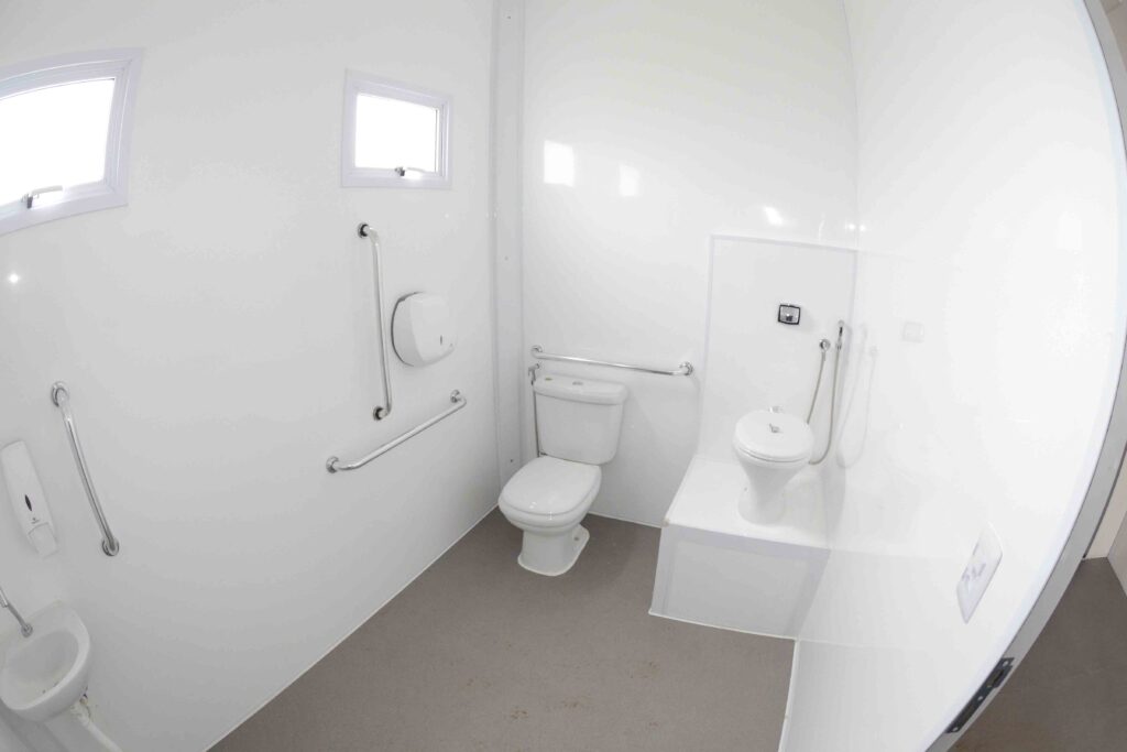 Esta imagem apresenta o interior moderno e limpo de um banheiro acessível, com paredes brancas brilhantes, piso cinza e instalações sanitárias adaptadas, criando um ambiente funcional, organizado e iluminado. A composição é capturada através de uma lente grande-angular que amplia o espaço reduzido, mostrando vários elementos essenciais dispostos estrategicamente para acessibilidade. No centro, há um vaso sanitário branco padrão, enquanto à direita encontra-se um vaso sanitário mais baixo ou infantil, instalado sobre uma plataforma elevada. Barras de apoio metálicas prateadas estão fixadas nas paredes ao redor dos sanitários e na parede esquerda para garantir a segurança dos usuários. Pequenas janelas retangulares estão posicionadas na parte superior das paredes esquerda e frontal, permitindo a entrada de luz natural. Um pequeno lavatório branco é visível no canto inferior esquerdo, complementado por um dispenser de sabonete líquido e um suporte para papel higiênico montado na parede. Os principais objetos são os dois vasos sanitários brancos, as barras de apoio em aço inoxidável e os acessórios sanitários, todos limpos e bem conservados. O vaso principal possui uma caixa acoplada na parte superior, enquanto o vaso menor na plataforma elevada está equipado com uma ducha higiênica flexível prateada. As barras de apoio possuem superfícies metálicas polidas e estão firmemente parafusadas nas paredes brancas. O piso cinza escuro contrasta com a luminosidade das paredes e do teto, proporcionando aderência e facilitando a limpeza do ambiente. A imagem é uma fotografia digital tirada com uma lente panorâmica ou olho de peixe, resultando em uma perspectiva ligeiramente curva nas bordas que abrange a totalidade do pequeno espaço. A execução técnica foca na clareza e na documentação visual, destacando a acessibilidade e o design funcional do local. Não há elementos artísticos estilizados, sendo um registro puramente utilitário e arquitetônico. O ambiente interno é iluminado por uma luz difusa e brilhante, possivelmente combinando a luz natural vinda das pequenas janelas superiores com iluminação artificial embutida, criando uma atmosfera asséptica, limpa e segura. As superfícies brancas refletem a luz, maximizando a sensação de claridade no espaço restrito. O clima geral é prático e higiênico, priorizando a utilidade, a segurança e o conforto para pessoas com necessidades especiais.