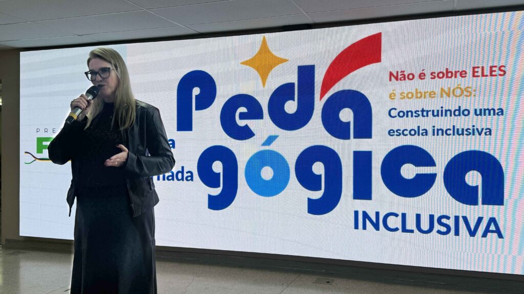 A imagem mostra uma mulher falando ao microfone diante de um grande painel digital com o título “Pedagógica Inclusiva”. O texto no painel diz: “Não é sobre ELES, é sobre NÓS: Construindo uma escola inclusiva”, indicando o tema de uma apresentação voltada à educação inclusiva.