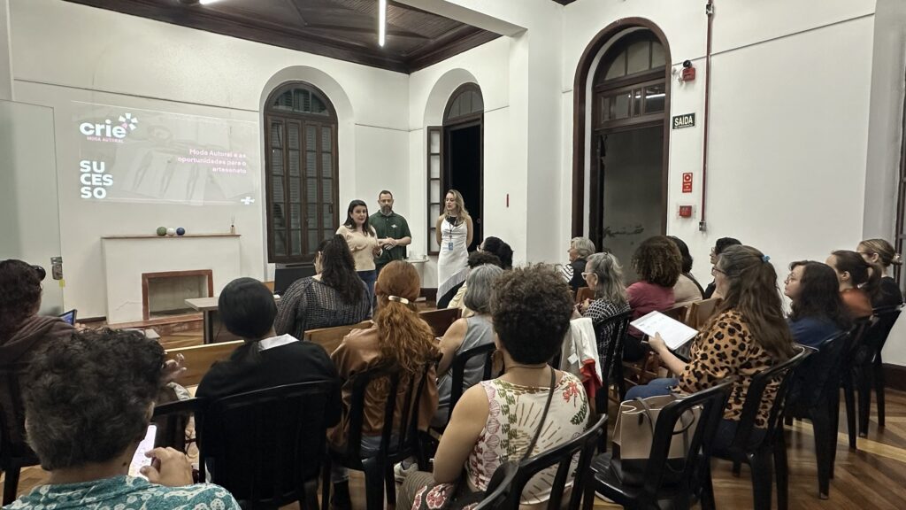 A imagem mostra uma reunião ou palestra em uma sala ampla, com várias pessoas sentadas em cadeiras e voltadas para três pessoas que estão à frente. Uma mulher parece estar falando ao grupo, acompanhada por um homem e outra mulher. Na parede, há uma projeção com o logotipo “crie Moda Autoral” e os textos “Moda Autoral e as oportunidades para o artesanato” e “SUCESSO”. O ambiente tem janelas e portas altas em arco, molduras de madeira escura, piso de madeira e uma lareira decorativa. Também aparecem placas de “SAÍDA” e equipamentos de emergência.