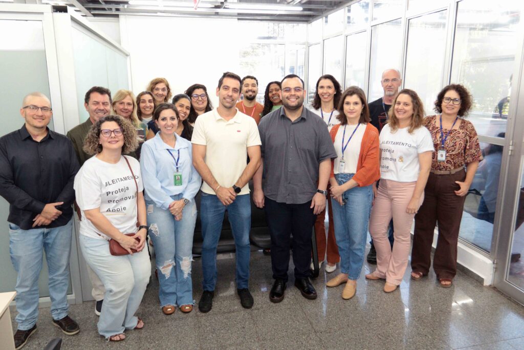 A imagem mostra uma foto de grupo em um ambiente interno, provavelmente uma sala de treinamento ou evento. Cerca de vinte pessoas aparecem sorrindo e posando juntas, muitas usando crachás e camisetas brancas com a palavra “ALEITAMENTO” e o nome “Promova”. Ao fundo há divisórias de vidro, cadeiras e uma parede clara. A foto está na orientação horizontal, mas foi enviada girada, e algumas pessoas nas bordas aparecem parcialmente cortadas.