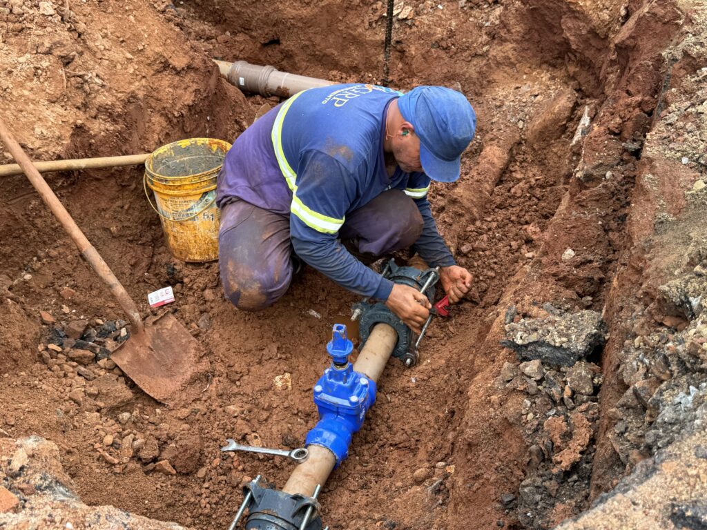 A imagem mostra uma obra de manutenção em uma vala de terra avermelhada. Um trabalhador, usando uniforme azul com faixas refletivas, está agachado ao lado de uma tubulação clara e de conexões azuis de PVC. No centro, há uma ferramenta elétrica amarela apoiada sobre o solo, além de uma pá ou cabo de madeira. Outro trabalhador aparece parcialmente no canto inferior direito, manuseando ferramentas junto à tubulação. A área está úmida e enlameada, com pedras, terra revolvida e água acumulada ao redor das conexões.