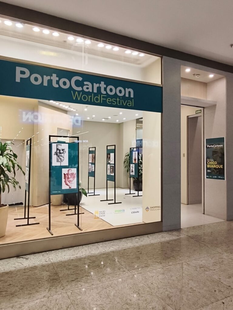 A imagem mostra uma galeria de arte com o letreiro “PortoCartoon World Festival”, evento internacional dedicado à caricatura e ao humor gráfico.