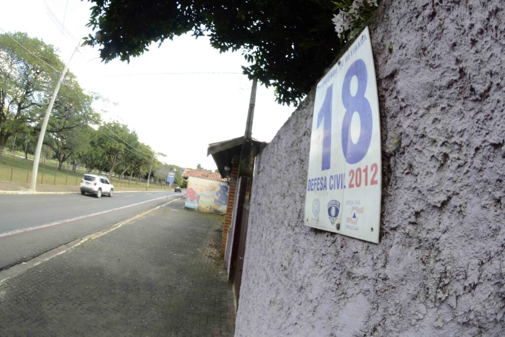 A imagem está inclinada e mostra uma rua arborizada vista ao lado de um muro de pedra. Há uma calçada de blocos, uma via com alguns carros e, do outro lado, uma área verde cercada, com árvores e gramado. No muro, aparecem cartazes coloridos parcialmente visíveis, com textos e ilustrações. Ao fundo, há uma pequena construção com telhado de cerâmica.