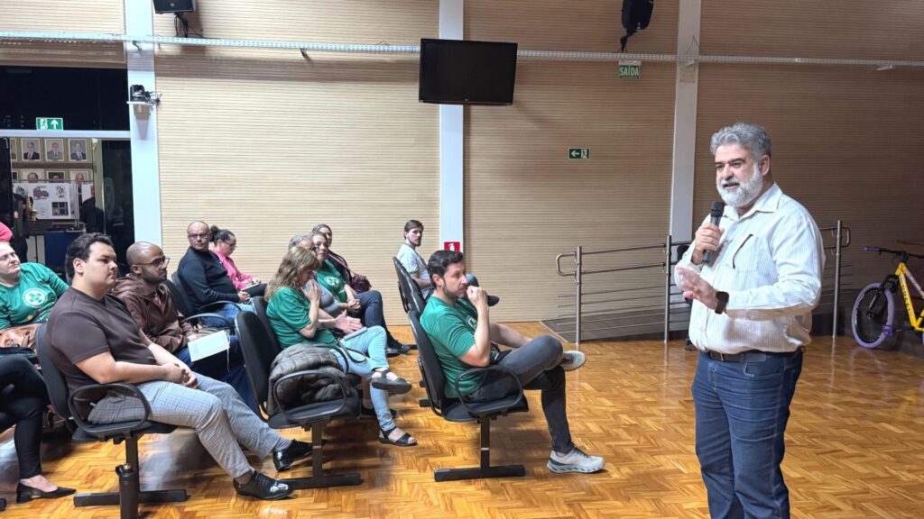 A imagem mostra uma reunião ou palestra em um auditório. Um homem de barba grisalha, vestindo camisa branca, está em pé com um microfone, falando para o público. Os participantes estão sentados em cadeiras, atentos à apresentação; alguns vestem camisetas verdes com um logotipo circular, sugerindo que pertencem a uma mesma organização ou grupo.