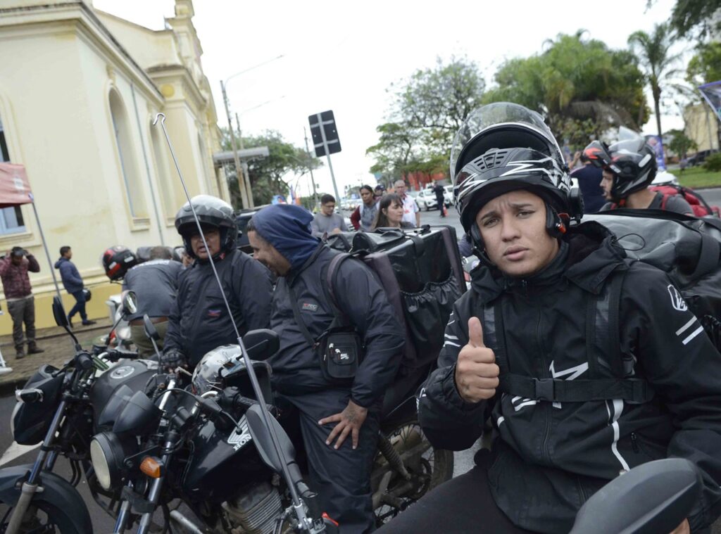 Um jovem estafeta/motoboy montado numa motocicleta faz um gesto de aprovação com o polegar para cima. Ele usa capacete e equipamento preto de proteção, acompanhado por outros motociclistas numa via pública. Na frente da motocicleta ao seu lado, destaca-se uma antena de proteção corta-cerol.