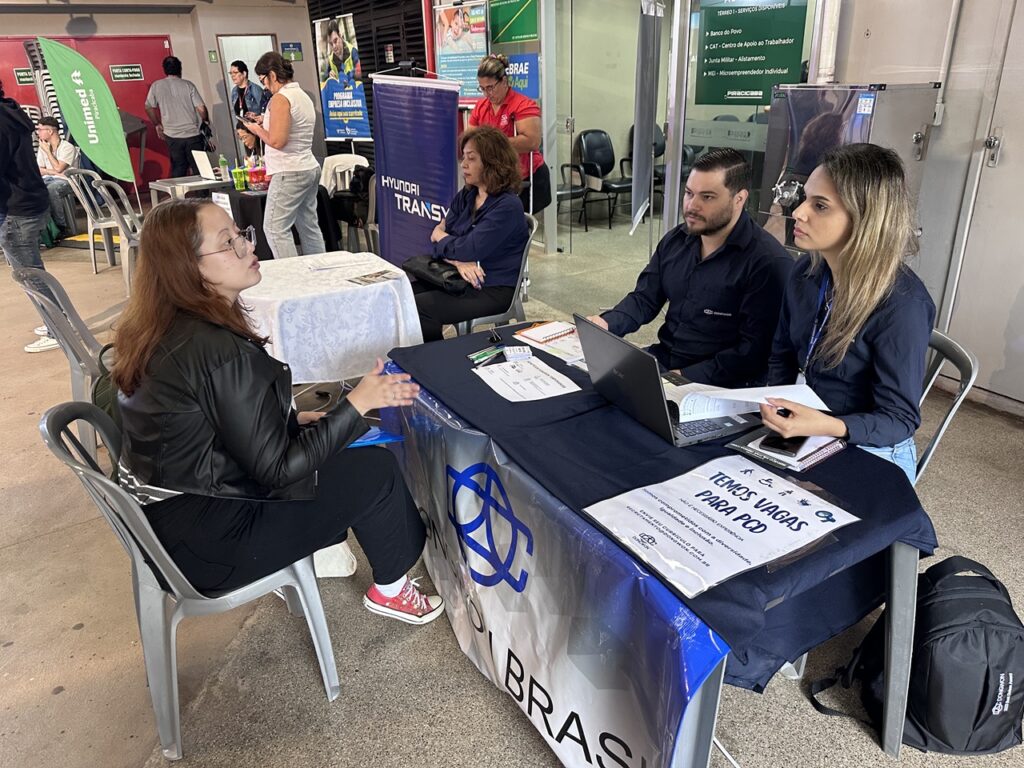 Participante conversa com representantes de uma empresa durante um feirão de empregos, em uma mesa de recrutamento com vagas disponíveis.