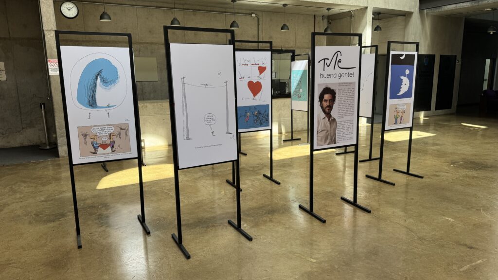 Exposição de cartuns e ilustrações em painéis verticais dispostos em um espaço cultural amplo, com obras gráficas e humorísticas.
