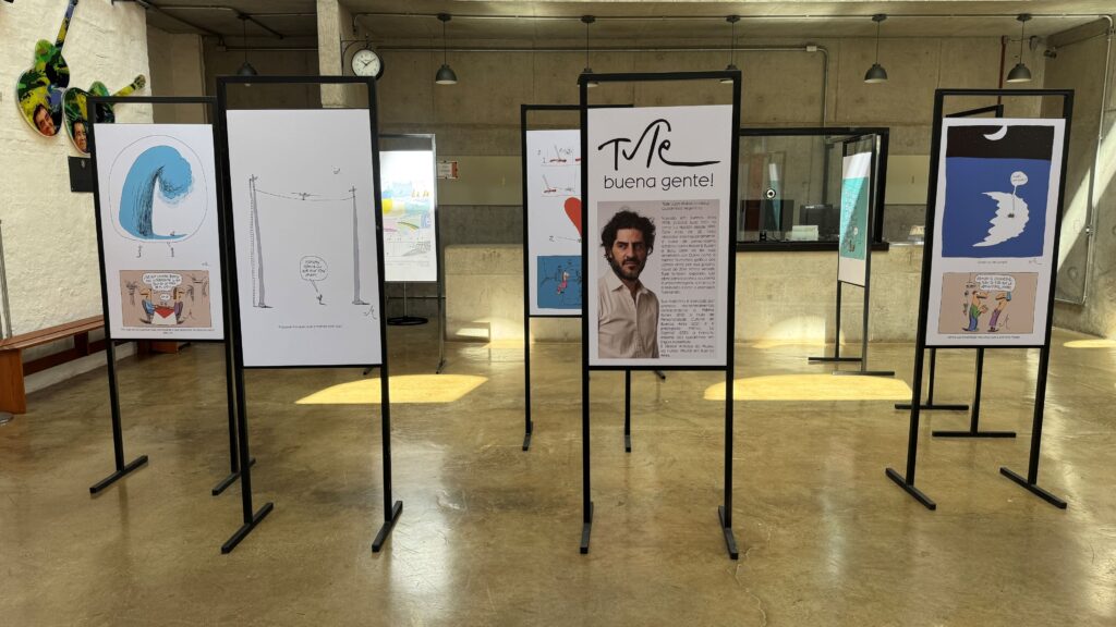 Mostra de ilustrações humorísticas e cartuns expostos em suportes metálicos em um ambiente cultural amplo e moderno.