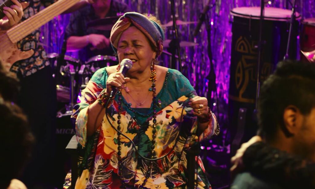 A imagem mostra uma apresentação musical ao vivo vibrante. No centro, uma mulher canta segurando um microfone, vestida com roupas coloridas e estampadas, adornada com colares e turbante. Ela parece profundamente envolvida na performance, com expressão de emoção e entrega.