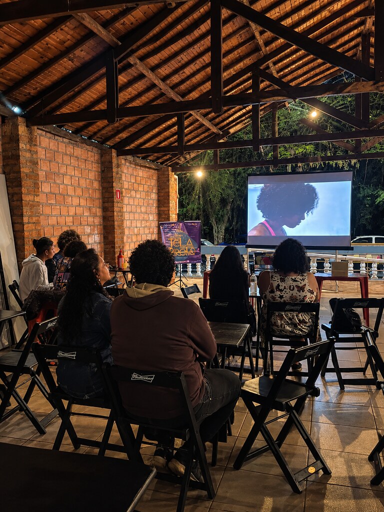 A imagem mostra um evento cultural em um espaço coberto com estrutura de madeira e paredes de tijolos aparentes. Um grupo de pessoas está sentado em cadeiras dobráveis, assistindo a uma projeção em tela grande que exibe uma mulher de perfil com cabelo crespo.
