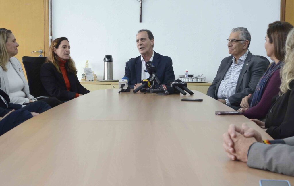 Nesta imagem, vemos uma cena de coletiva de imprensa ou anúncio oficial em uma sala de reuniões. Um homem, posicionado ao centro da mesa, fala diante de vários microfones de diferentes veículos de mídia, reforçando o caráter público e formal do momento. Ao redor, outras pessoas acompanham atentamente, vestidas em trajes de negócios, o que transmite profissionalismo. No fundo, há elementos simples como um pequeno crucifixo na parede, uma garrafa térmica de café e uma bandeja com xícaras, que acrescentam um toque cotidiano ao ambiente institucional.