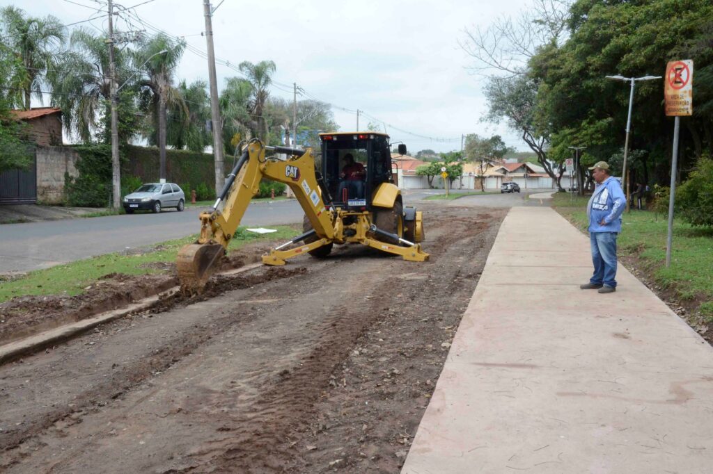 A imagem mostra uma retroescavadeira amarela da marca CAT realizando escavação ou nivelamento de solo ao lado de uma calçada recém‑construída.