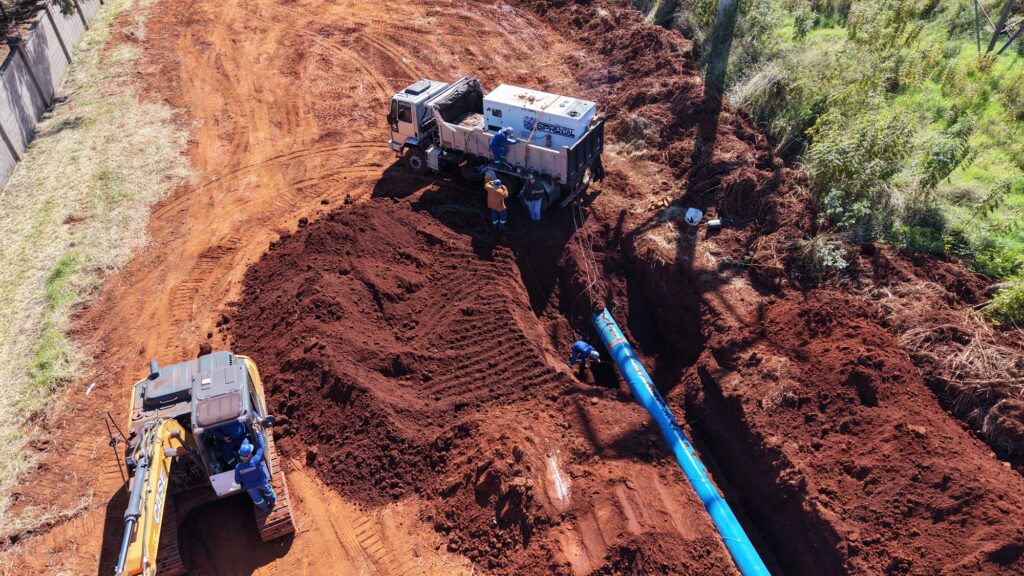 A construção está em andamento em uma área com terra vermelha rica em minerais, com algumas gramíneas verdes e uma cerca de concreto no lado esquerdo da imagem. A terra recém-escavada forma uma paisagem acidentada, marcada por sulcos profundos e montes de terra. Um caminhão branco com uma unidade geradora no seu compartimento de carga e o nome "SPRENTAL" escrito nas laterais está estacionado numa área mais elevada de terra. Um grupo de quatro trabalhadores, todos vestindo roupas de trabalho de cores vibrantes, parece estar a interagir com a carga do camião. Dois deles estão dentro da caixa do camião, um em pé e outro agachado, enquanto dois estão fora, perto da porta traseira do camião. Um dos trabalhadores fora está com um macacão laranja e calças azuis e botas, enquanto o outro usa calças azuis e um camisola cinzenta. Há um outro trabalhador na parte de trás do camião que está a descer para o solo, vestindo um macacão azul. Um escavador amarelo da marca "CASE" está posicionado na parte inferior esquerda da imagem, com um trabalhador vestindo um macacão azul ao lado dele, perto da cabine. Parece que o escavador acabou de mover a terra vermelha que domina a paisagem. No lado direito da imagem, uma longa tubagem azul repousa numa vala escavada. Um trabalhador, vestido com um macacão azul, está a trabalhar na zona onde a tubagem é colocada. Há também uma quantidade de vegetação verde e arbustos que crescem à volta da zona de construção, indicando um ambiente mais natural nas imediações. A composição da imagem é dominada pela terra vermelha, que ocupa a maior parte do espaço, criando uma sensação de trabalho em curso. A luz do sol ilumina a cena, projetando sombras distintas que realçam as texturas da terra. A paleta de cores é dominada pelos tons de terra vermelha, com o branco do camião e o azul da tubagem e das roupas de trabalho a contrastarem e a chamarem a atenção para as atividades humanas. A perspectiva aérea proporciona uma visão abrangente do local de construção e da tarefa que está a ser realizada.