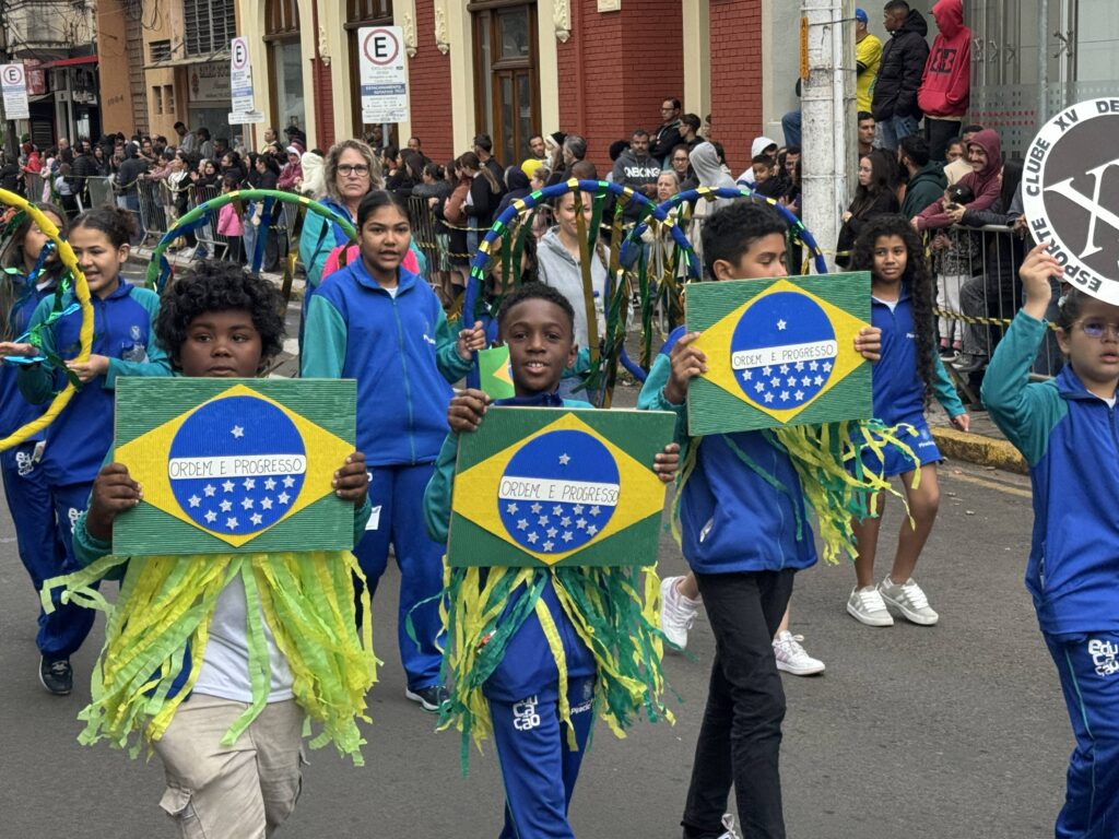 Na imagem, vemos um grupo de crianças participando de um desfile cívico pelas ruas da cidade, vestidas em roupas azul e verde. Elas carregam placas retangulares com a bandeira do Brasil e o lema “Ordem e Progresso”, algumas decoradas com fitas verdes e amarelas, reforçando o tom patriótico da celebração. Ao fundo, há prédios, placas de rua e espectadores acompanhando atrás das grades, compondo uma atmosfera festiva e comunitária que valoriza a identidade nacional e o espírito coletivo.