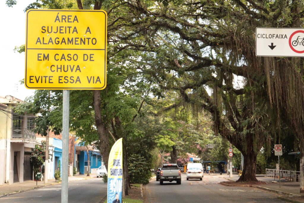 A imagem mostra uma via urbana cercada por árvores grandes e frondosas, criando um ambiente sombreado e agradável. Em destaque, há uma placa amarela de alerta com os dizeres: “Área sujeita a alagamento. Em caso de chuva, evite essa via.”