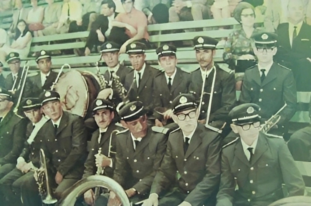 A imagem parece ser uma fotografia antiga, com tonalidade esverdeada, mostrando uma banda ou corporação musical uniformizada. Os músicos usam casacas escuras, gravatas e quepes com emblemas; alguns seguram saxofones, trombones e outros instrumentos de sopro, enquanto um grande bumbo aparece ao centro. Eles estão reunidos em arquibancadas, diante de um público sentado. A foto tem aparência envelhecida e apresenta algumas áreas desfocadas.