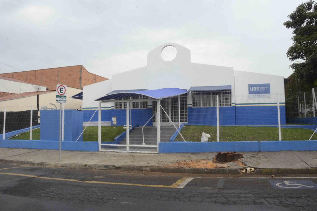 Uma fotografia ao nível dos olhos mostra a fachada de um posto de saúde pública, uma UBS, em um dia nublado, com um prédio branco e azul cercado por uma grade metálica branca e localizado em uma rua asfaltada. A composição é simétrica e frontal, centralizando a fachada principal do edifício de saúde. A estrutura é dividida horizontalmente entre paredes brancas na parte superior e detalhes em azul escuro e claro na base e nas faixas decorativas. Na frente, há um pequeno gramado ladeando uma escadaria de acesso central que leva à entrada principal, protegida por um pequeno teto curvo de lona azul. Uma cerca metálica branca delimita a propriedade, com um portão fechado no centro, alinhado com a escadaria. À esquerda, uma placa de trânsito de proibido estacionar ergue-se na calçada cinza de concreto. O edifício principal apresenta uma arquitetura simples e funcional, caracterizada por paredes pintadas em branco na metade superior e um tom azul vibrante na metade inferior. Acima da entrada, a fachada eleva-se em um frontão triangular que exibe uma abertura circular vazada, lembrando um óculo. Janelas com grades metálicas brancas estão dispostas de forma simétrica em ambos os lados da entrada. Na lateral direita, uma placa azul indica a sigla "UBS" junto com o logotipo do governo local. A entrada possui portas e janelas de vidro protegidas por grades de segurança. A imagem é uma fotografia digital documental colorida, tirada com luz natural difusa sob um céu nublado, o que elimina sombras fortes e produz uma iluminação suave e uniforme em toda a cena. A perspectiva frontal enfatiza a clareza arquitetônica do edifício institucional. A nitidez é consistente em toda a imagem, registrando detalhes como a textura do asfalto, o concreto da calçada e os tijolos expostos de construções vizinhas ao fundo. O cenário urbano apresenta uma rua asfaltada escura em primeiro plano, com marcações de sinalização horizontal parcialmente visíveis e um símbolo de vaga para deficientes pintado no canto inferior direito. A calçada é limpa, feita de blocos de cimento cinza. O ambiente é calmo e deserto, sem a presença de pedestres ou veículos em movimento. Ao fundo, estruturas de tijolos aparentes de construções adjacentes e árvores com folhagens verdes escuras completam o horizonte urbano, criando uma atmosfera sóbria, institucional e cotidiana.