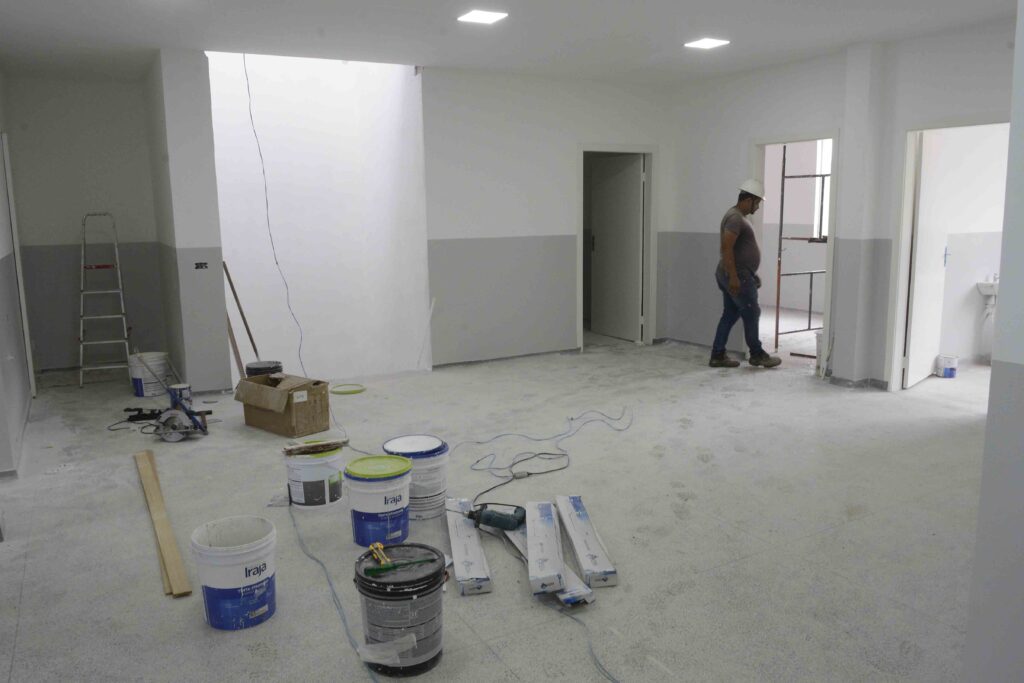 Uma fotografia realista retrata o interior de uma ampla sala em plena reforma, onde um trabalhador realiza suas tarefas cercado por ferramentas, baldes de tinta e materiais de construção espalhados pelo chão de cimento. A composição da cena é ampla e aberta, mostrando um espaço interno em processo de acabamento. À esquerda, há uma escada de alumínio apoiada na parede e diversos baldes e caixas organizados desordenadamente. No centro, o chão exibe fios elétricos soltos, ferramentas manuais e embalagens de materiais. À direita, um trabalhador caminha em direção a uma das portas, enquanto outras aberturas revelam cômodos adjacentes. A distribuição dos elementos cria uma sensação de atividade em andamento, típica de um canteiro de obras interno. O principal sujeito humano é um trabalhador de construção civil que veste uma camisa cinza escura, calça jeans e um capacete de segurança branco. Ele aparece em perfil, caminhando com postura concentrada, carregando equipamentos ou inspecionando o local. Pelo chão, destacam-se vários baldes de tinta plásticos com rótulos coloridos, ferramentas elétricas como uma serra circular e uma furadeira, além de caixas de papelão e tábuas de madeira dispostas aleatoriamente. A imagem é uma fotografia digital documental que registra com clareza e realismo um ambiente real de trabalho. A execução técnica foca na nitidez e na captura de luz natural e artificial, sem filtros estilizados, priorizando a fidelidade visual dos materiais de construção, das texturas das paredes e do piso empoeirado. O cenário se passa em um ambiente interno fechado, possivelmente um escritório ou uma residência em reforma. A iluminação é proveniente de luminárias quadradas embutidas no teto branco e de aberturas estruturais que deixam entrar luz difusa, criando uma atmosfera neutra, funcional e típica de um espaço em transformação.