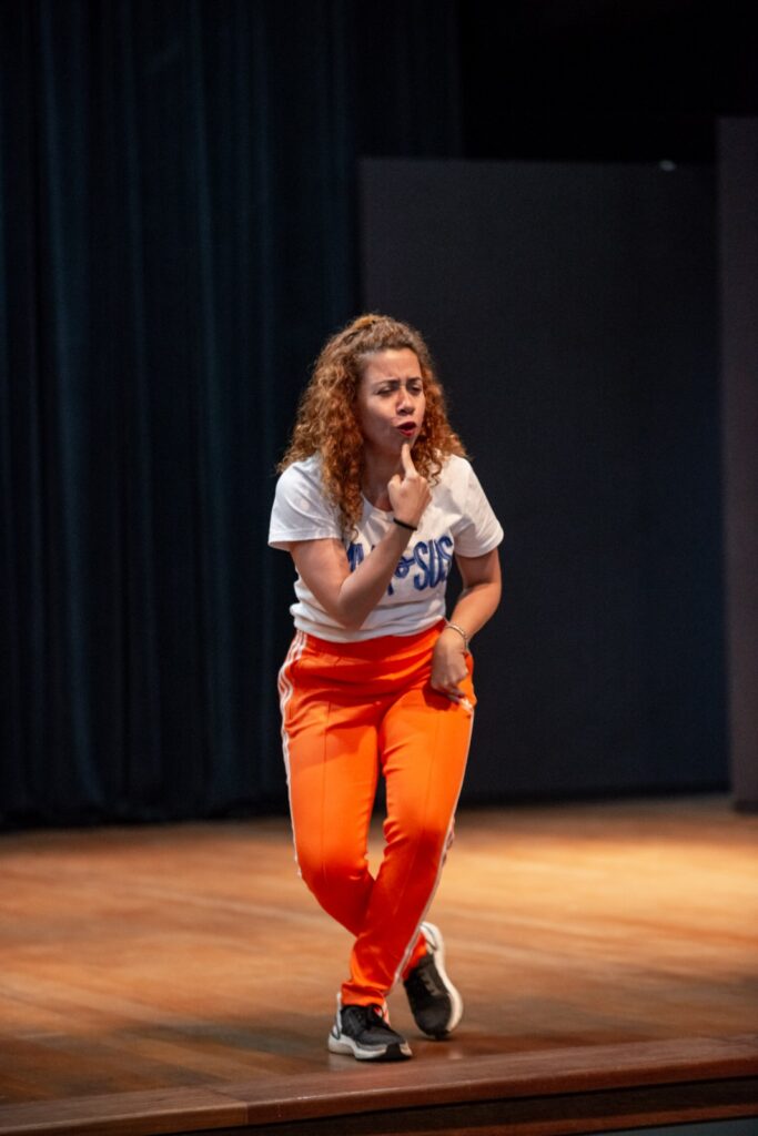 A imagem mostra uma pessoa em um palco com fundo escuro, usando camiseta branca e calça laranja