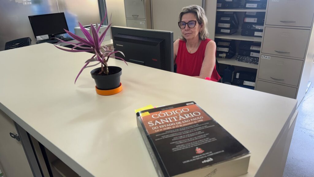 Ambiente de escritório com um balcão amplo. Ao fundo, uma pessoa está sentada diante de um computador. Sobre o balcão há um vaso com planta de folhas roxas e um livro intitulado “Código Sanitário do Estado de São Paulo”. Armários e pastas organizadas aparecem ao fundo.