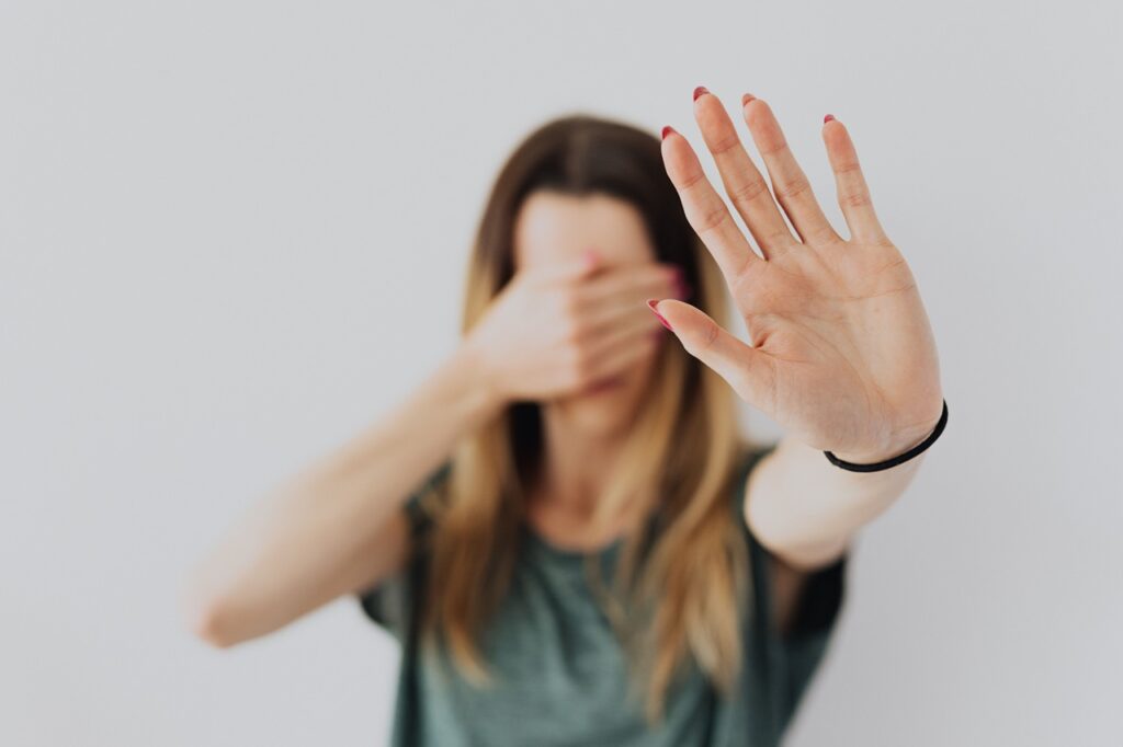 A imagem mostra uma mulher com cabelos castanhos claros, vestindo uma camiseta verde, em frente a um fundo branco neutro. Ela está com uma mão estendida em direção à câmera, com a palma aberta — gesto que pode indicar recusa, bloqueio ou pedido de distância — enquanto cobre o rosto com a outra mão.