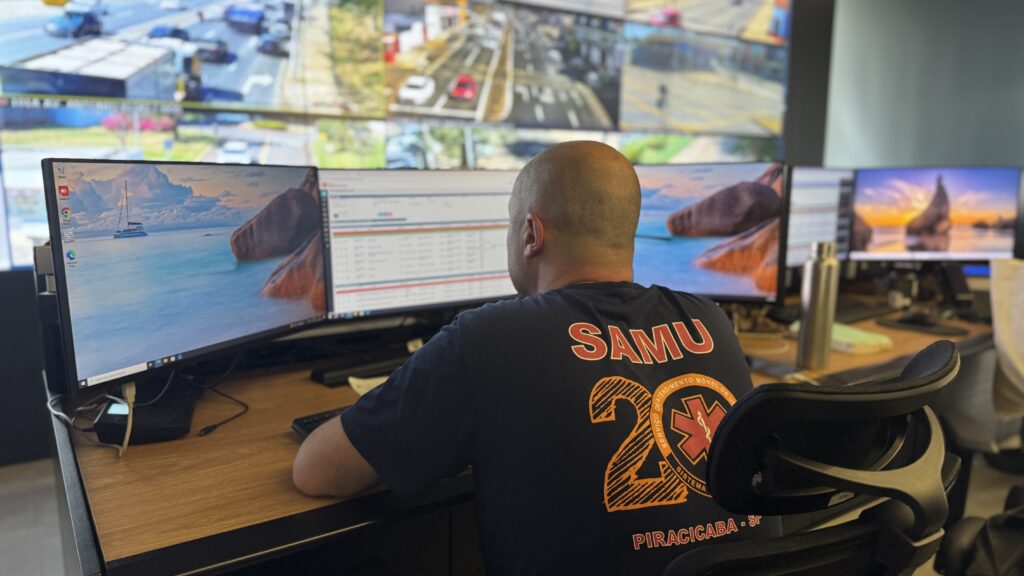 A imagem mostra uma pessoa sentada diante de vários monitores de computador, em um ambiente de trabalho ou central de atendimento. As telas exibem páginas da internet e imagens de paisagens, enquanto a pessoa usa uma camiseta preta com estampas e textos em laranja, incluindo “Piracicaba”. A cena está fotografada de lado e com pouca nitidez, destacando o perfil da pessoa, os monitores e a mesa de trabalho.