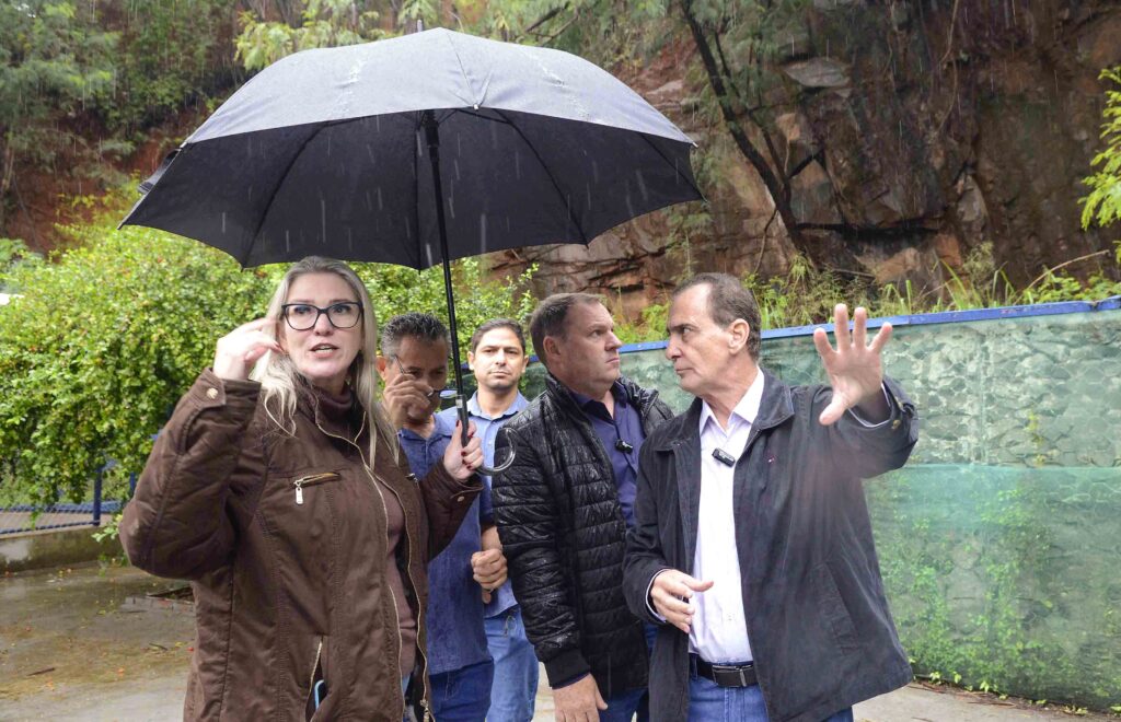 Quatro pessoas estão reunidas em uma área externa, sob chuva, ao lado de um paredão de rocha. Um homem gesticula enquanto conversa com o grupo, protegido por um guarda-chuva.