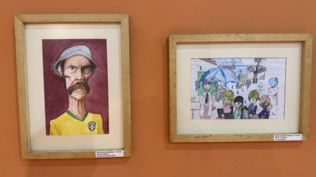 Dois quadros em caixilho de madeira pendurados numa parede alaranjada: à esquerda, uma caricatura da personagem Seu Madruga com a camisola da seleção brasileira de futebol; à direita, um desenho infantil a ilustrar uma sala de aula durante a chuva.