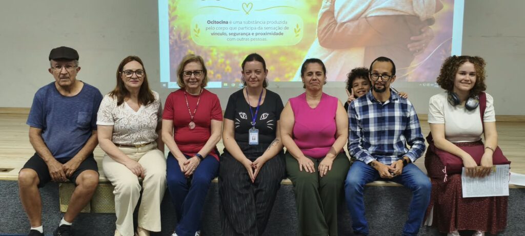 Sete pessoas estão sentadas lado a lado num auditório, em frente a uma tela de projeção sobre ocitocina. Uma criança espreita de forma divertida por trás do grupo.
