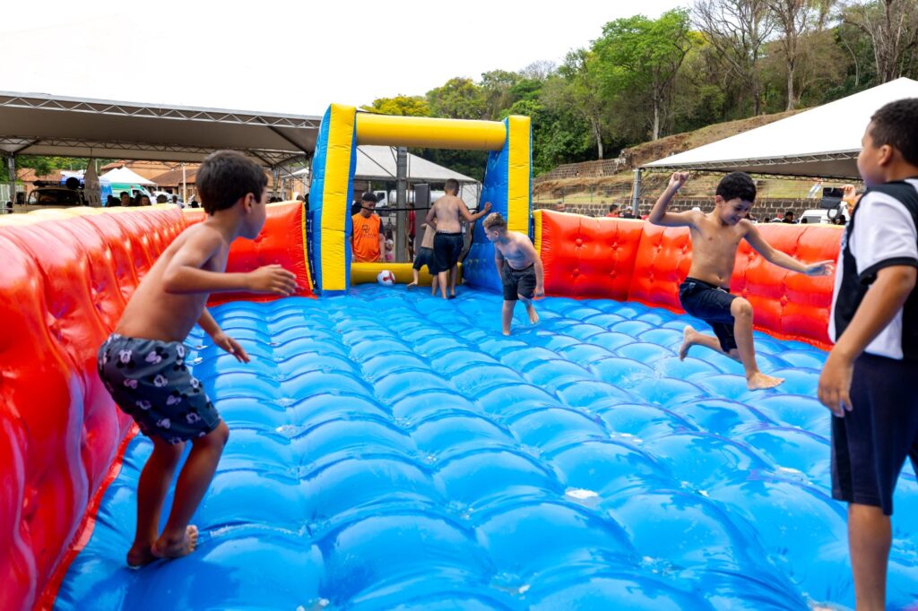 Crianças a brincar e a saltar num campo de futebol insuflável gigante com chão azul e bordas vermelhas.