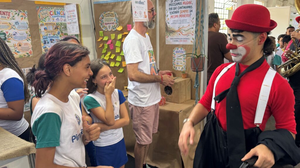 Um palhaço caracterizado com chapéu e nariz vermelho interage e diverte duas alunas nos corredores da feira.