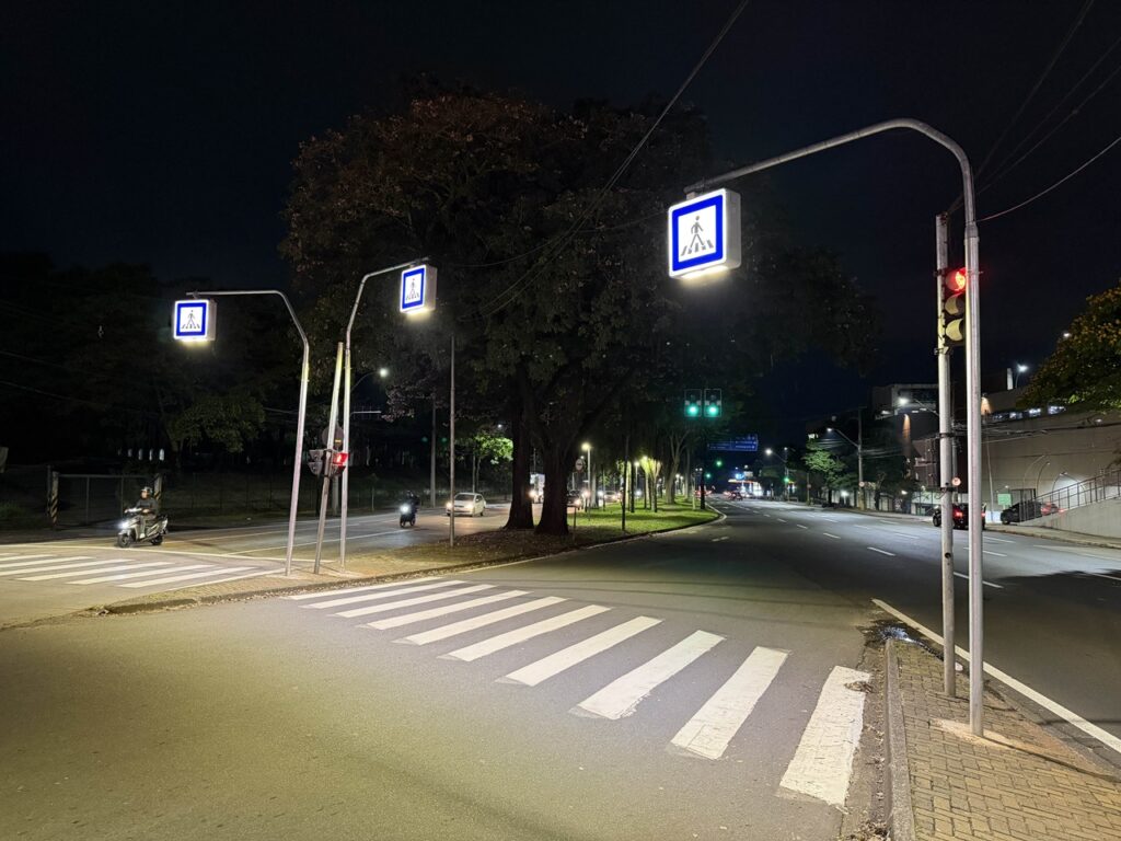 A imagem mostra uma vista noturna de uma avenida urbana bem iluminada, destacando uma passagem de peões no primeiro plano
