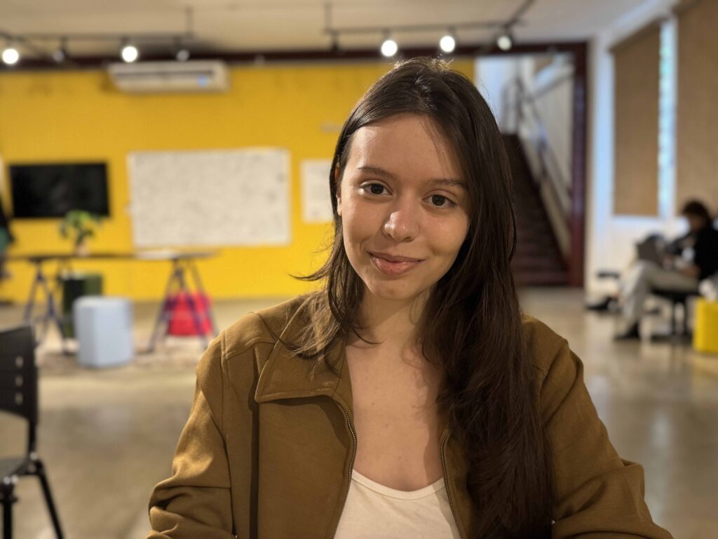 Retrato em plano médio da artista plástica Yasmim Agostinho, vestindo um casaco acastanhado e sorrindo num espaço amplo iluminado ao fundo.