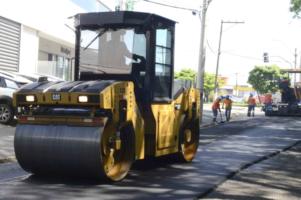Plano aproximado de um grande rolo compactador amarelo em movimento sobre a camada fresca de asfalto. Ao fundo, trabalhadores acompanham a pavimentação da via ao lado de outros equipamentos de obras.