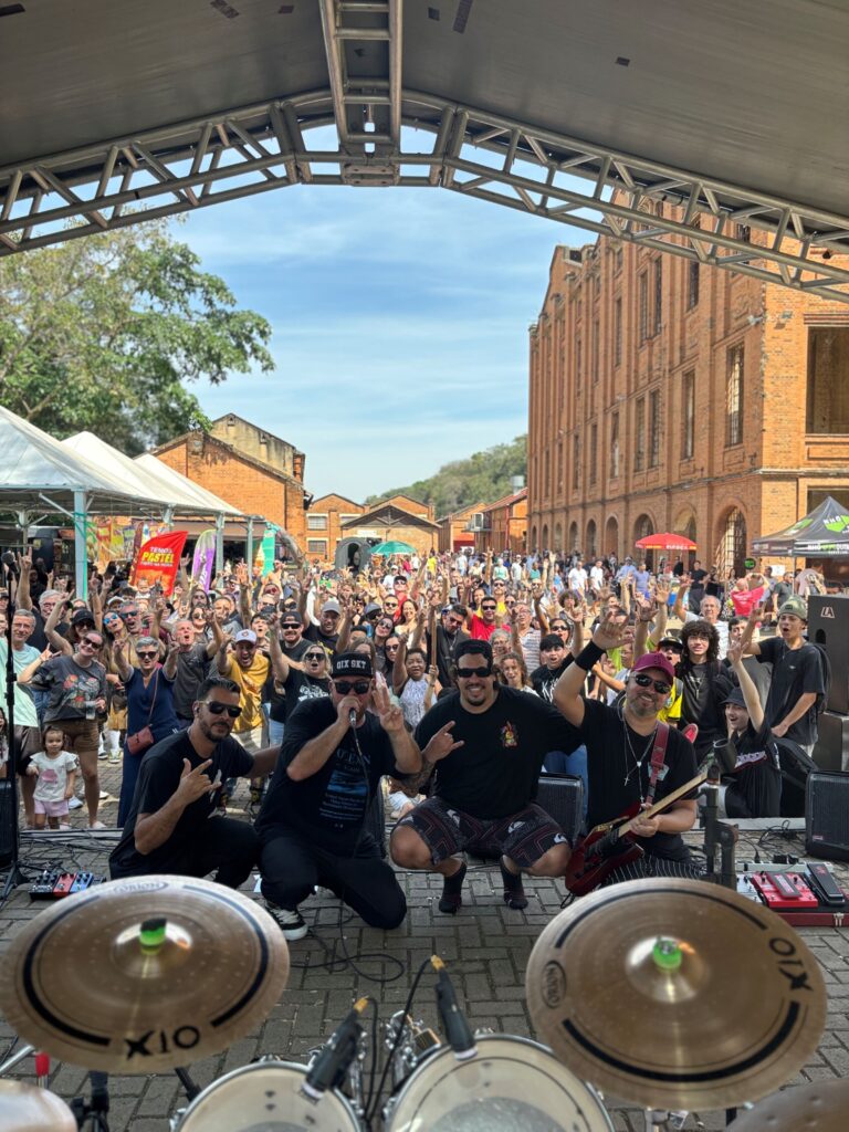Show de uma banda em um evento ao ar livre. Os músicos aparecem no palco, em frente a uma grande plateia reunida entre construções de tijolos aparentes. O público está voltado para o palco, com várias pessoas levantando os braços.