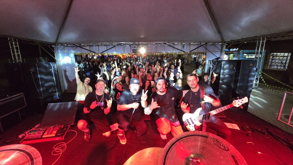 Apresentação musical noturna sob uma tenda. A banda posa no palco diante de uma multidão, com iluminação vermelha destacando os músicos. O público ocupa a área em frente ao palco, acompanhando o show.