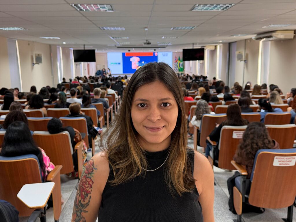 Retrato em plano médio da coordenadora Sangela Nascimento, com o auditório lotado de participantes ao fundo.
