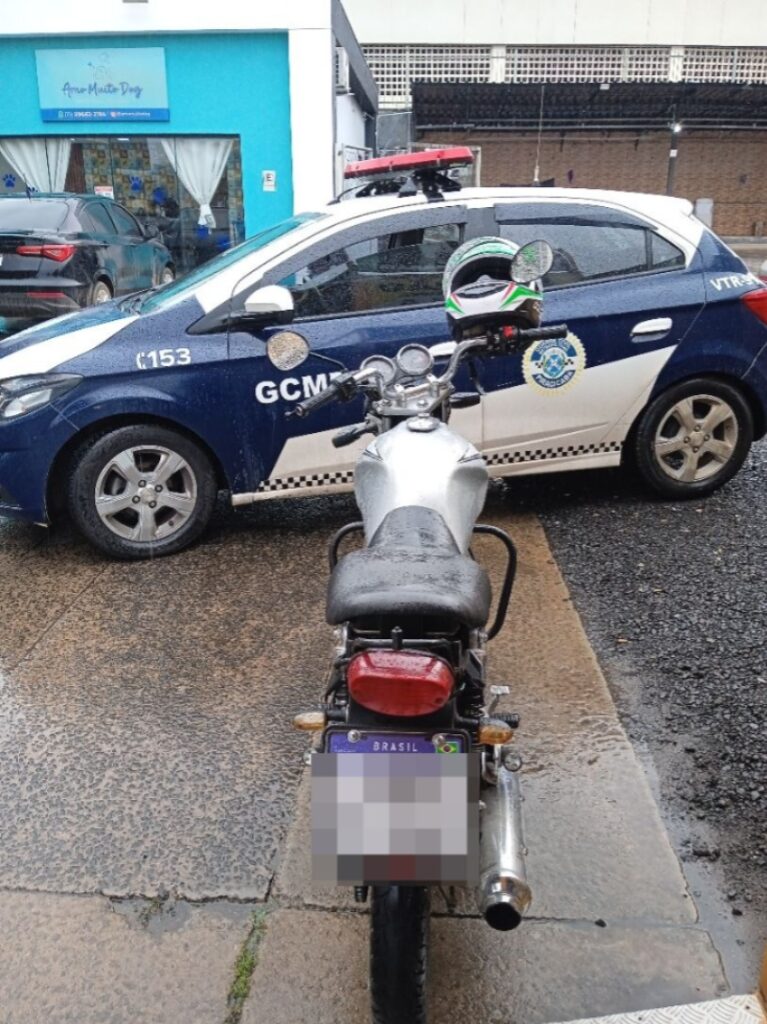Uma motocicleta prateada estacionada num pátio molhado pela chuva, mesmo em frente a um carro patrulha da Guarda Civil Municipal de Piracicaba.