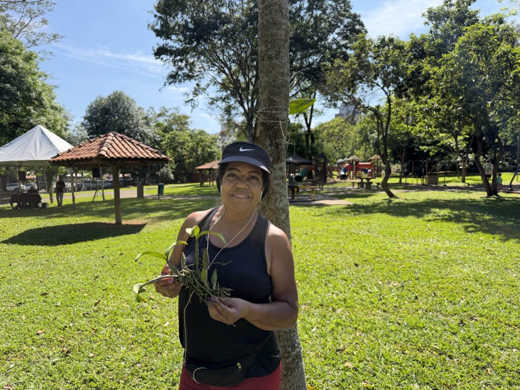 Retrato de uma mulher a sorrir, com viseira e roupa desportiva, de pé junto a uma árvore. Ela segura com as duas mãos uma muda de orquídea antes de ser fixada. Ao fundo, avista-se o relvado do parque com quiosques de telha e parque infantil.