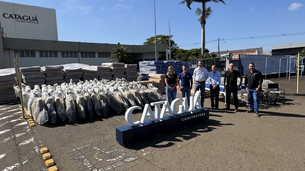 Vista em plano aberto do pátio do estacionamento com um grupo de seis pessoas alinhadas ao fundo, atrás do letreiro tridimensional "CATAGUÁ CONSTRUTORA". A grande quantidade de equipamentos hospitalares e colchões doados encontra-se disposta de forma organizada por todo o pátio.