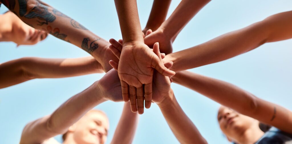 Mãos de diversas pessoas unidas no centro, sobrepostas umas sobre as outras num gesto de união e colaboração, filmadas a partir de um ângulo baixo contra um céu azul limpo.