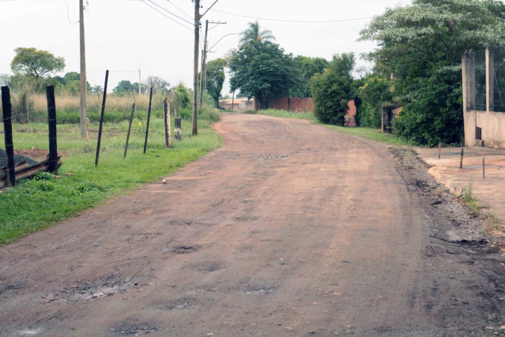 Estrada de terra e asfalto degradado numa área periférica ou rural, ladeada por cercas, postes de energia e árvores.