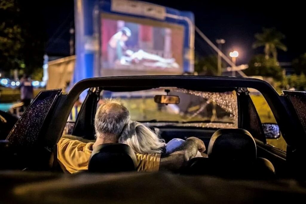 Perspetiva intimista a partir do interior de um veículo conversível. Um casal abraçado assiste ao filme projetado no ecrã gigante do cinema ao ar livre durante a noite.