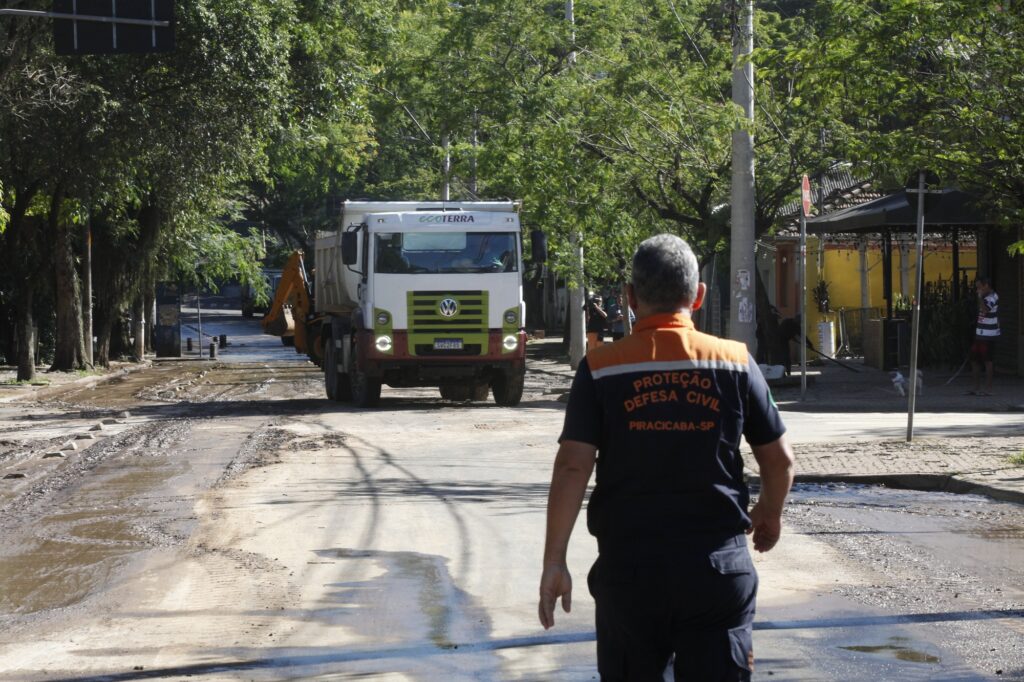 Regista um agente da Proteção e Defesa Civil de Piracicaba-SP de costas, vestido com o colete oficial, a supervisionar o tráfego e as operações na rua. Mais ao fundo, vê-se um caminhão de caçamba (EcoTerra) e uma máquina escavadora a operar na via coberta de lama.