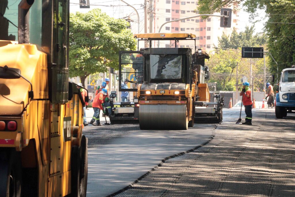 Mostra trabalhos de asfaltamento numa via urbana em progresso. Em destaque, veem-se rolos compactadores amarelos da marca Caterpillar e trabalhadores com coletes refletores e capacetes a operar o maquinário e a nivelar o asfalto.