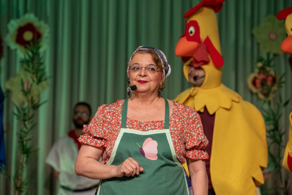 A maestrina e diretora Cíntia Pinotti, vestida com lenço na cabeça e avental com borboleta bordada, sorri em primeiro plano à frente de um ator fantasiado de galinha.